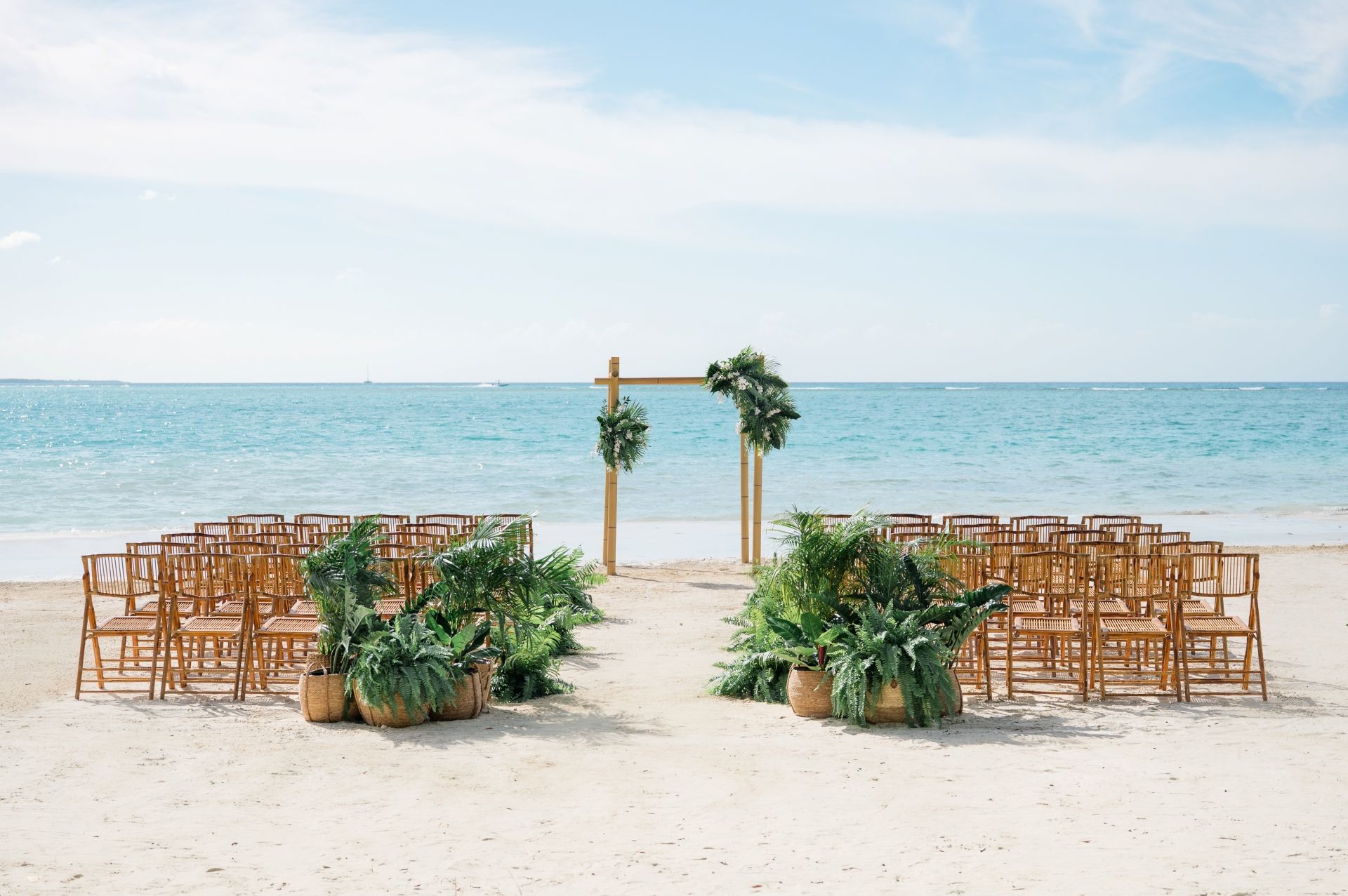 A tropical beach wedding ceremony setup with bamboo arch, lush greenery, and rattan chairs facing the turquoise Caribbean sea