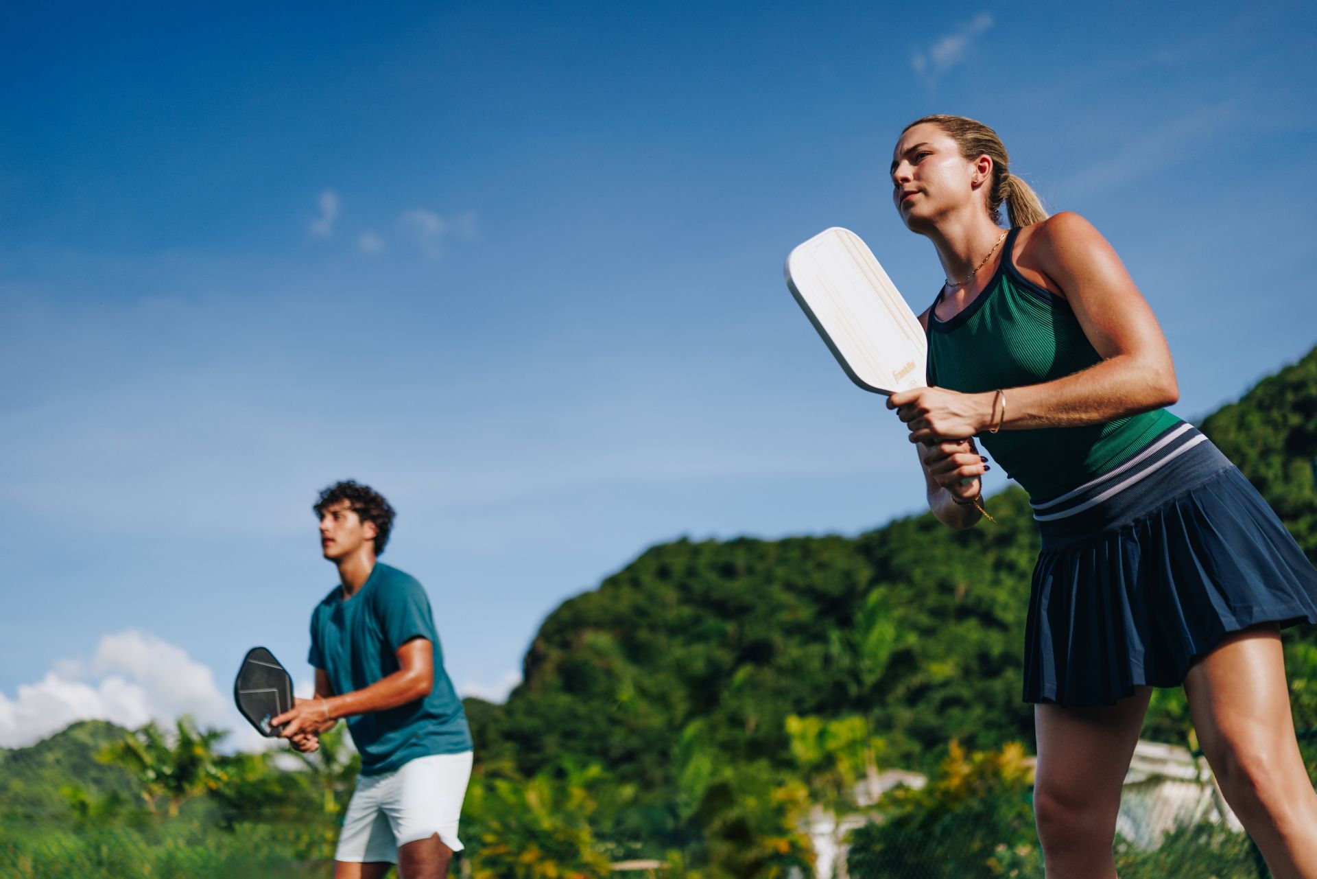 A couple plays pickleball on an outdoor court against a backdrop of lush green Caribbean mountains at Sandals Saint Vincent
